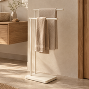 Toallero de pie de metal blanco roto con dos barras y toallas beige en baño de estilo mediterráneo