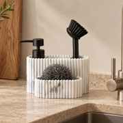 Organizador de fregadero efecto mármol blanco con dispensador de jabón negro, cepillo y estropajo metálico, sobre encimera de cocina beige.