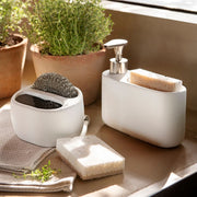 Organizador de fregadero blanco con dispensador de jabón, esponja y estropajo en ambiente de cocina.